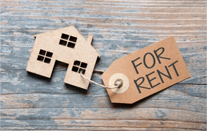 A small model house laying on a wooden surface with a paper tag attached with a piece of string reading "for rent"