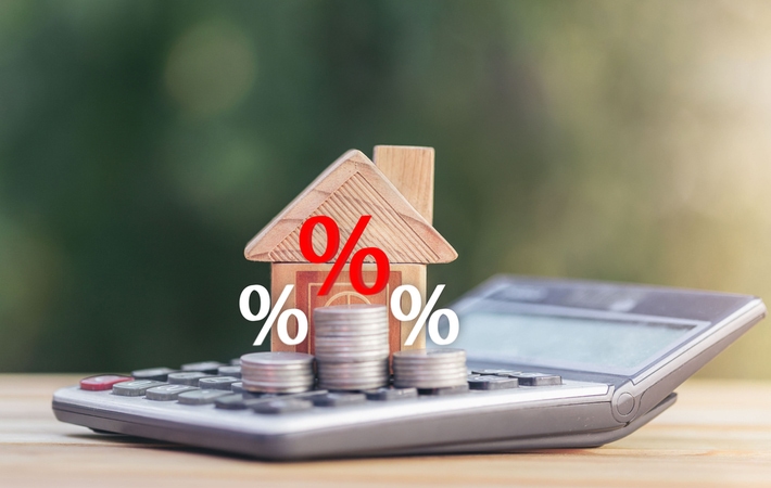 A small wooden house and 3 stacks of coins with percentage signs hovering above the coins, all resting on a calculator symbolizing mortgage interest rates