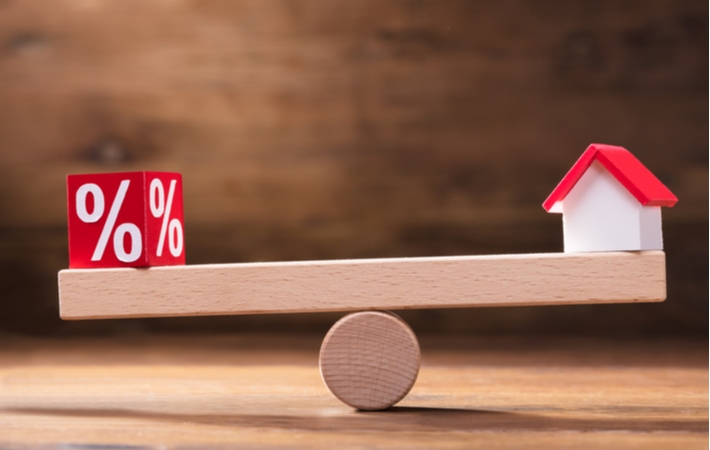 A wooden block with a percentage sign balancing on a small wooden seesaw across from a small wooden model house