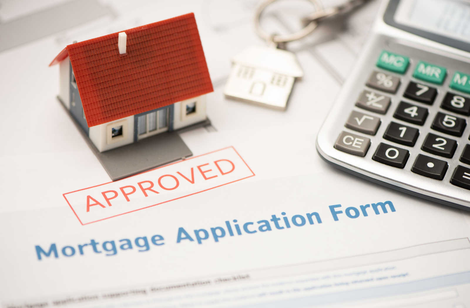 A calculator and model home sitting on top of a mortgage application form with "approved" stamped in red on top.