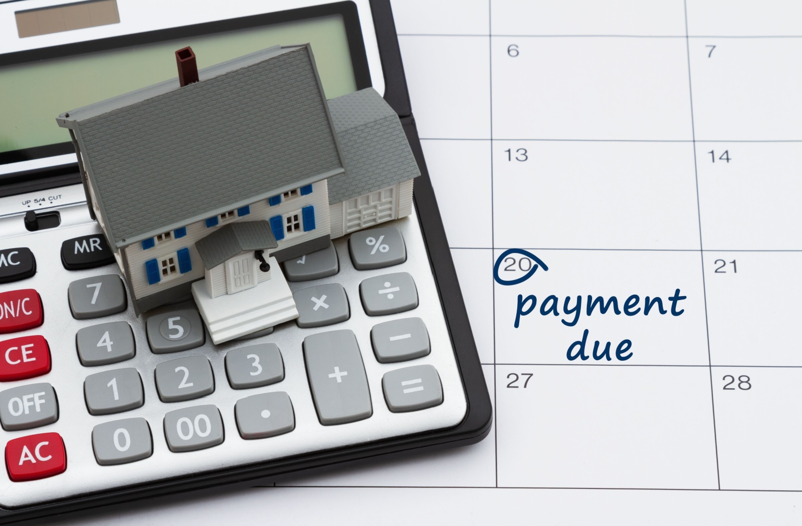A tiny model house resting on a calculator that sits on top of a calendar with the date circled to highlight a payment due date.