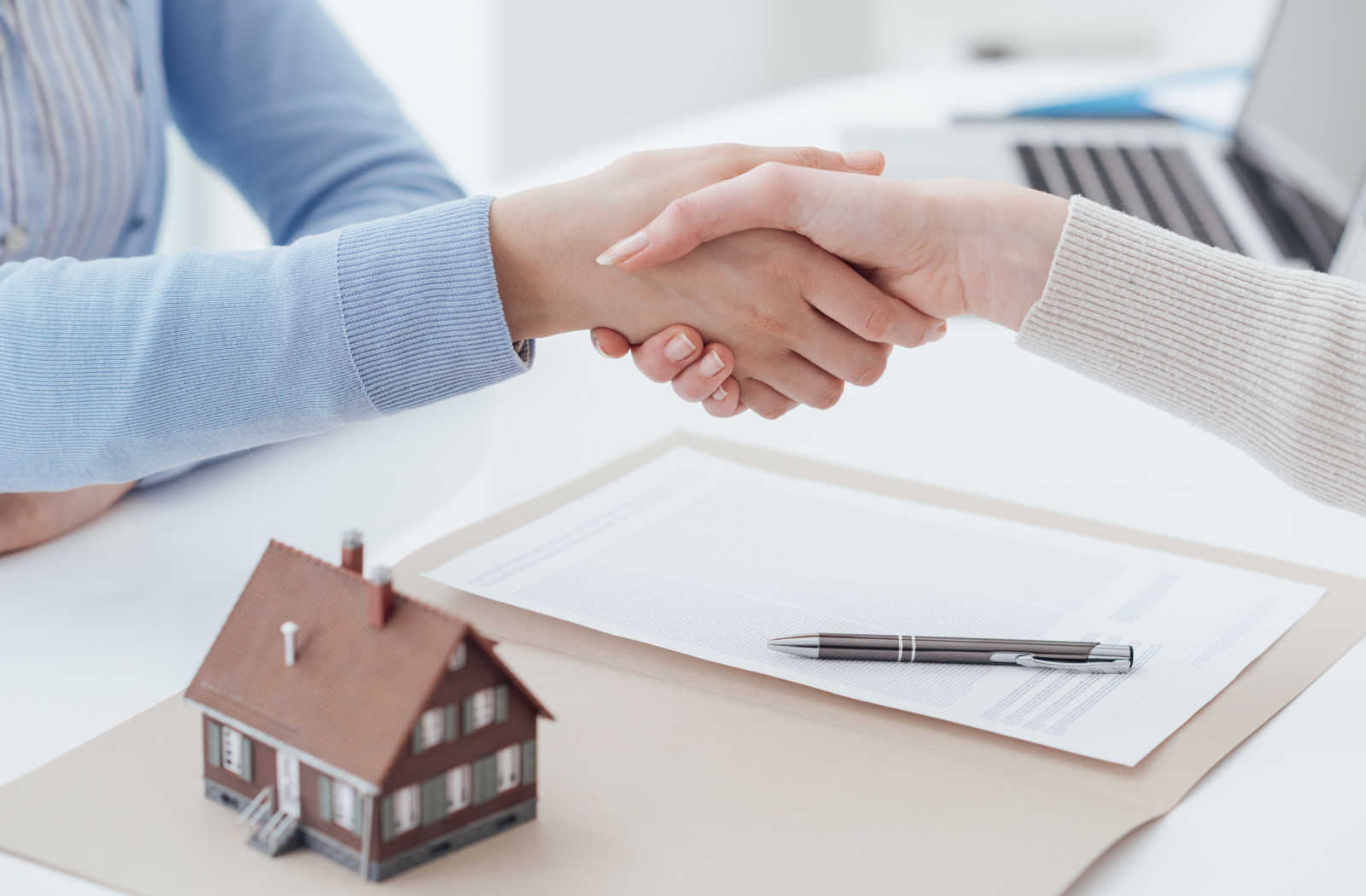 A client and mortgage broker shaking hands over a mortgage document with a model house sitting on the desk