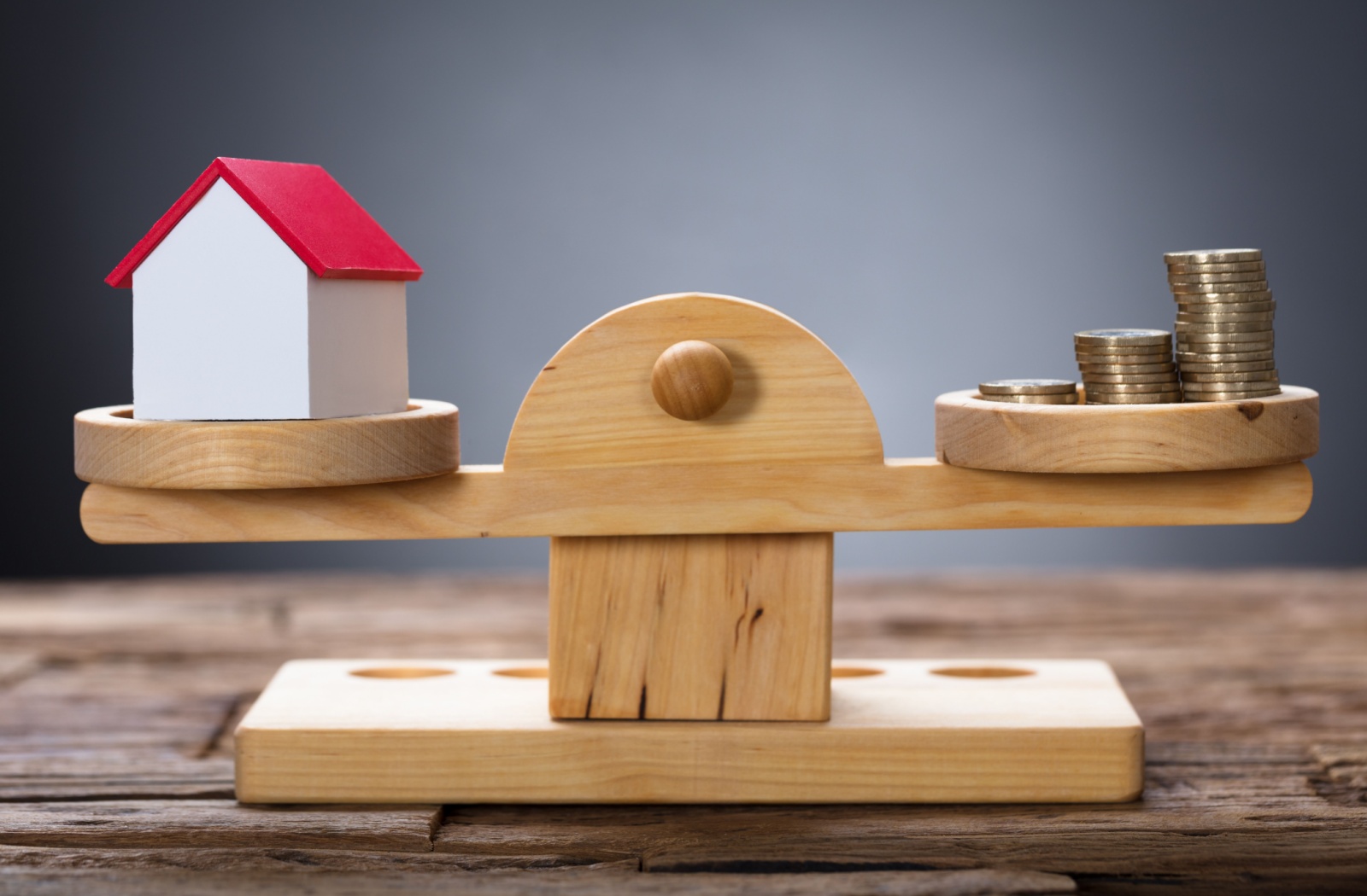 Closeup of model home and coins balancing on wooden weighing scale, representing a mortgage balance.