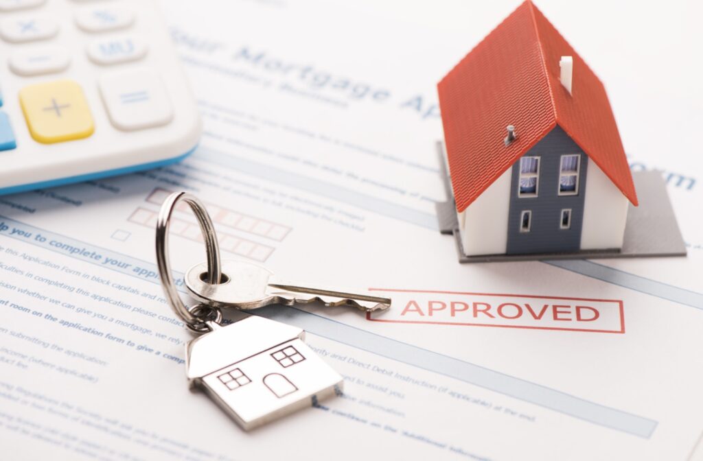 A mortgage application stamped with "approved" with keys and a model house sitting on top of the paper.