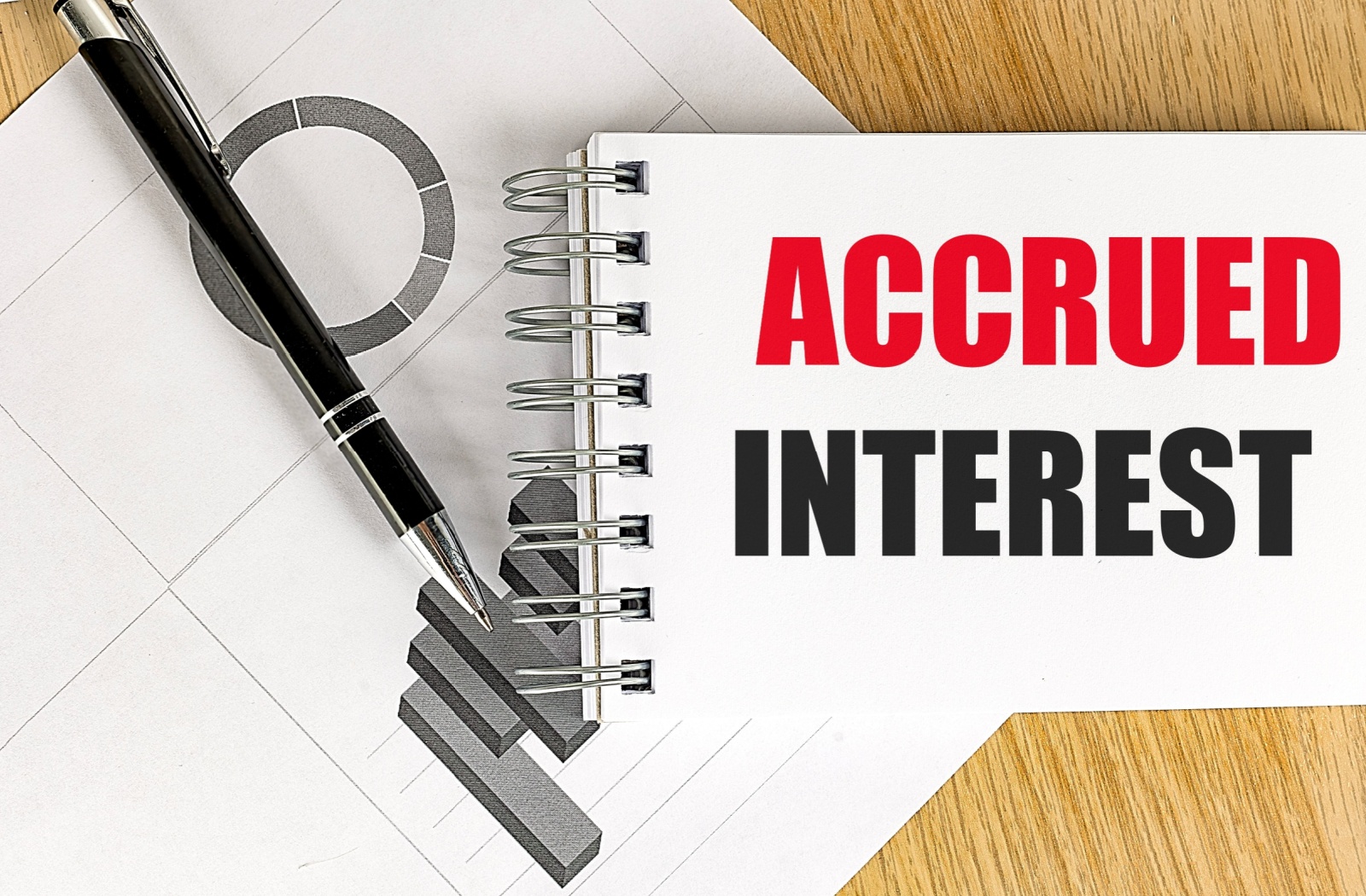 A notebook labeled "accrued interest", pen, and sheet with graphs on a table.